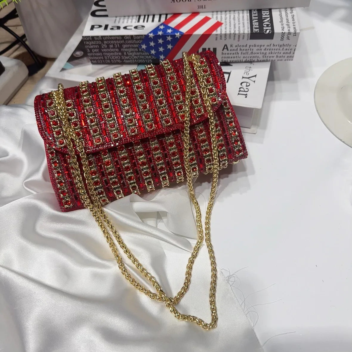 A red and gold rhinestone leather evening clutch bag lying on a white surface next to some books.