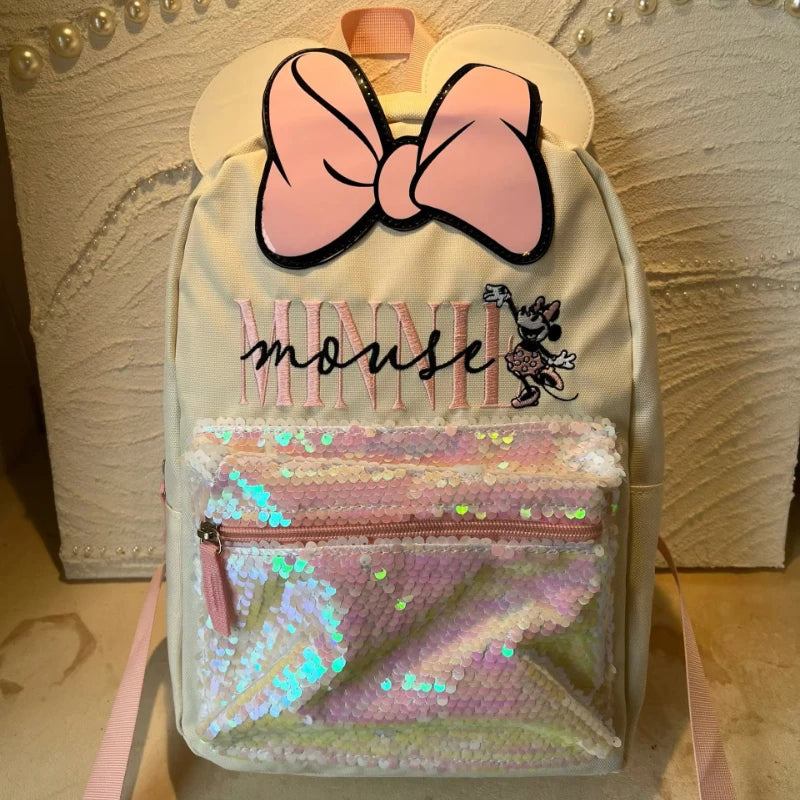 A light cream backpack decorated with a pink bow and a clear zippered pocket filled with sparkly pink and green sequins.