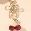 Pendentif clé en forme de nœud doré avec des pierres claires et un charm de cerise rouge pendu.