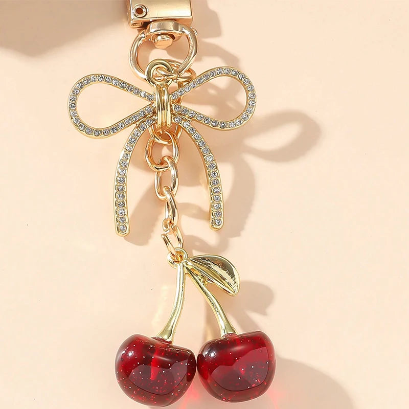 Pendentif clé en forme de nœud doré avec des pierres claires et un charm de cerise rouge pendu.
