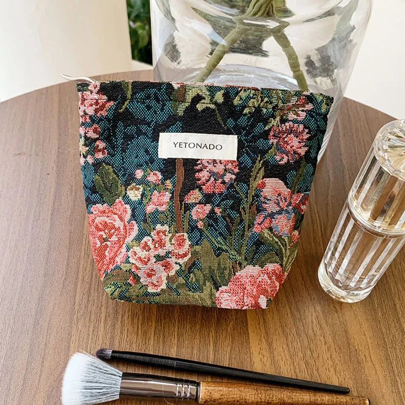 A dark floral fabric pouch with pink and green flowers is displayed next to a glass jar and a brush on a wooden surface.