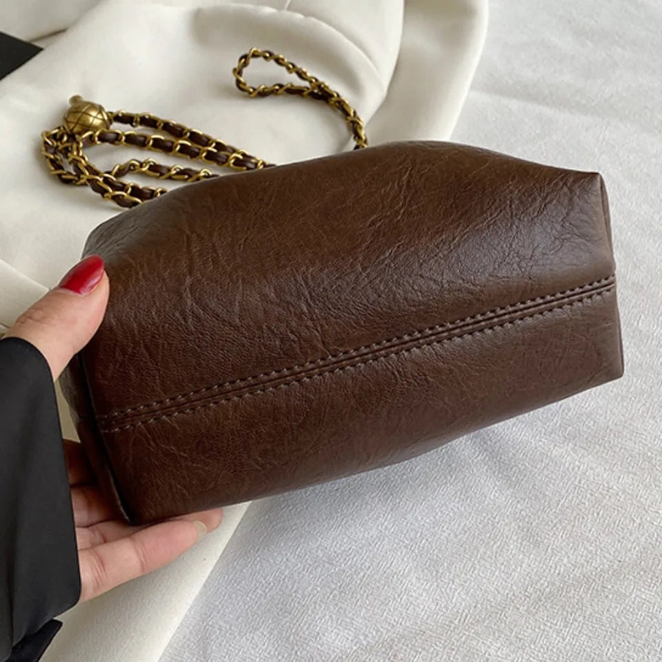A dark brown PU leather chain shoulder bag resting on a white fabric surface with a gold chain nearby.