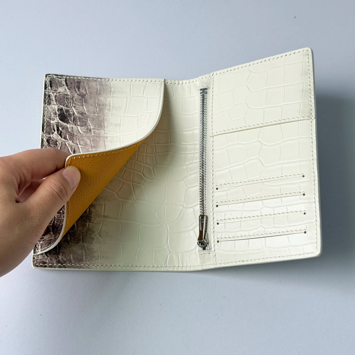 A close-up of a cream leather passport holder with a crocodile pattern and custom initials, shown being opened by a hand.