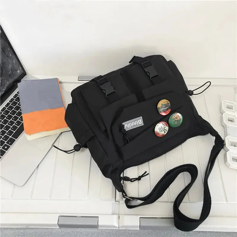 Una bolsa cruz tipo nylon negra casual mostrada sobre una superficie blanca junto a una laptop y un cuaderno.