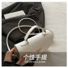 A white leather shoulder bag with gold hardware is shown resting on a dark gray fabric, highlighting its details.