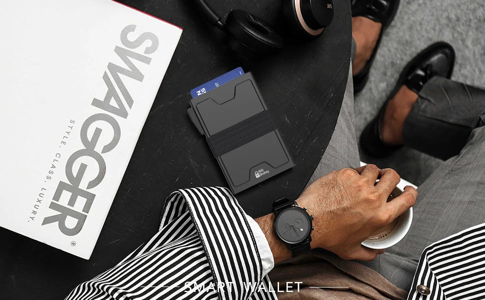A hand holds a minimalist metal card holder on a dark surface next to a magazine.