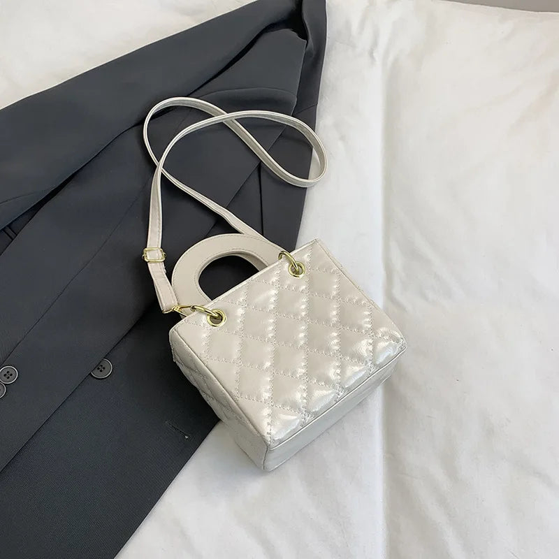 A light cream quilted crossbody handbag with a gold chain accent resting on a white surface next to a dark gray blazer.
