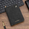 A black slim passport wallet with gold stitching and a silver airplane logo resting on a wooden surface next to a pen.