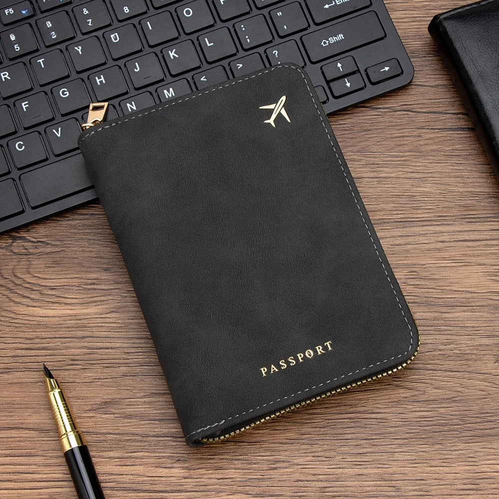 A black slim passport wallet with gold stitching and a silver airplane logo resting on a wooden surface next to a pen.
