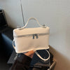 A cream-colored minimalist pebbled leather box crossbody bag with a silver zipper is held by a hand against a light backgroun