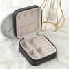 A black compact leather jewelry travel box with a lid open, resting on a white surface next to a clock.