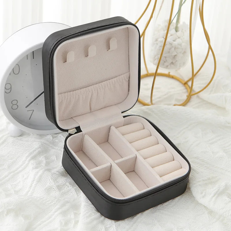 A black compact leather jewelry travel box with a lid open, resting on a white surface next to a clock.