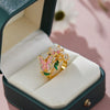 Gold rings featuring pink and green enamel butterfly designs displayed in a dark green jewelry box.