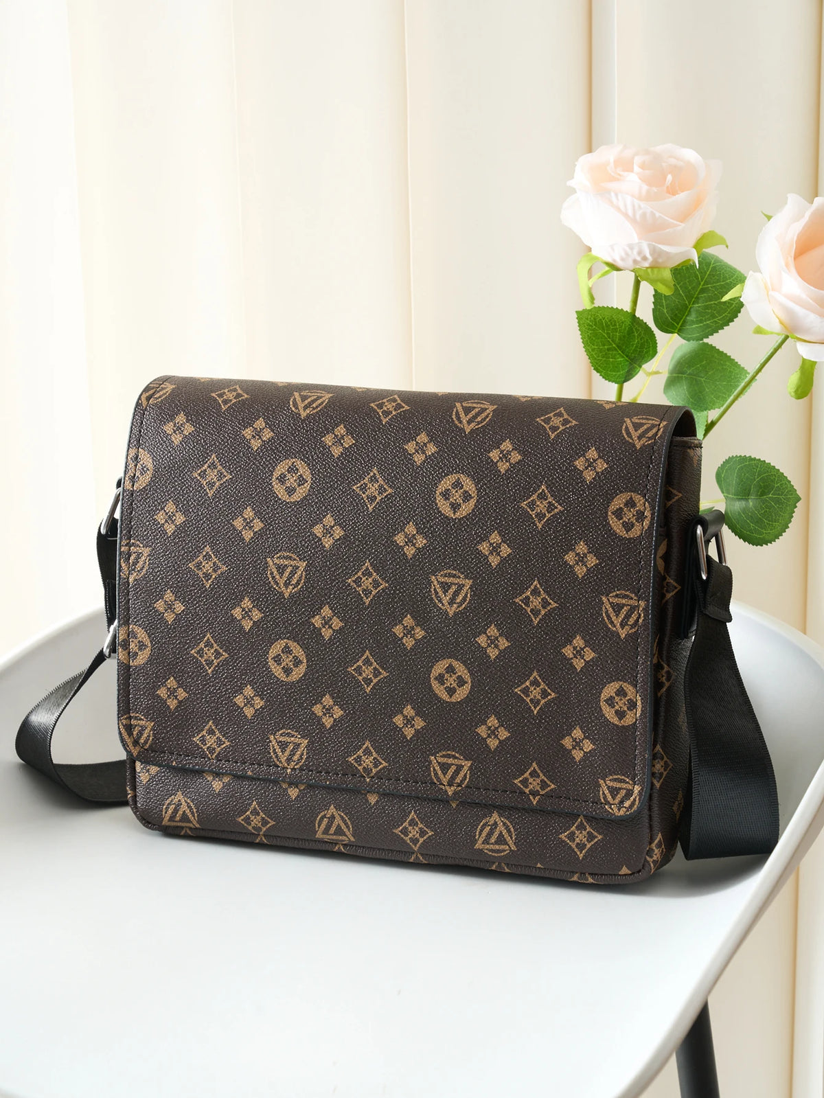 A brown men's casual crossbody bag with an easy flap design resting on a white surface next to pink roses.