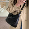 A woman wearing a beige trench coat holds a black PU leather tote bag with a strap and a white smartphone.