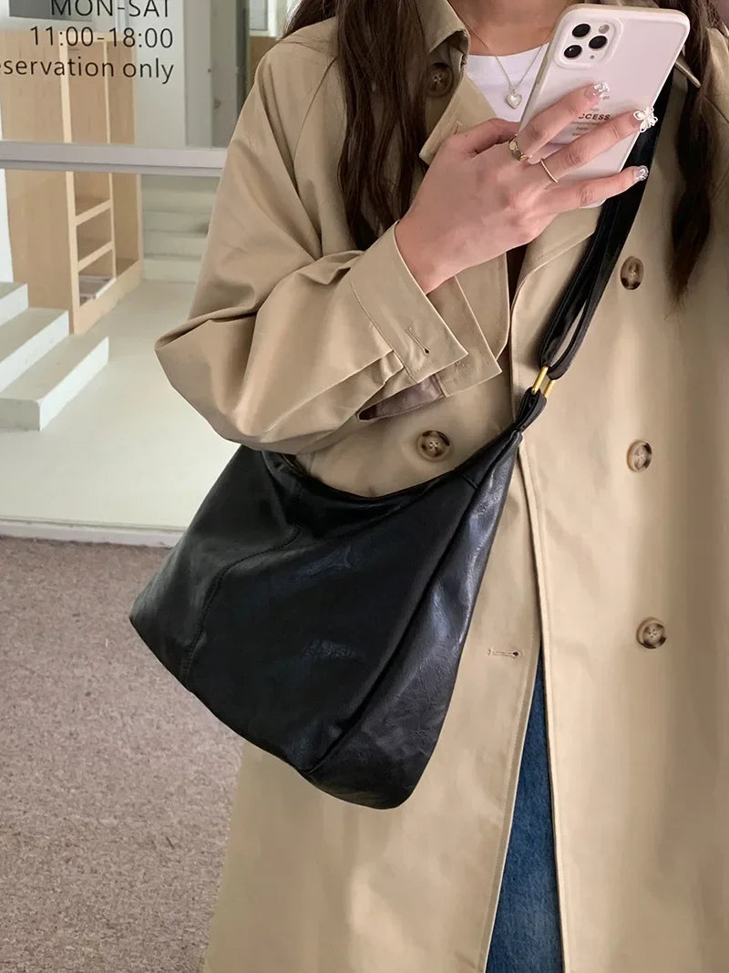 A woman wearing a beige trench coat holds a black PU leather tote bag with a strap and a white smartphone.