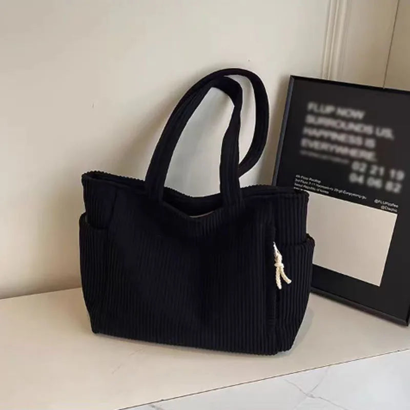 A black corduroy shoulder bag with a zipper is displayed on a white surface next to a framed sign.