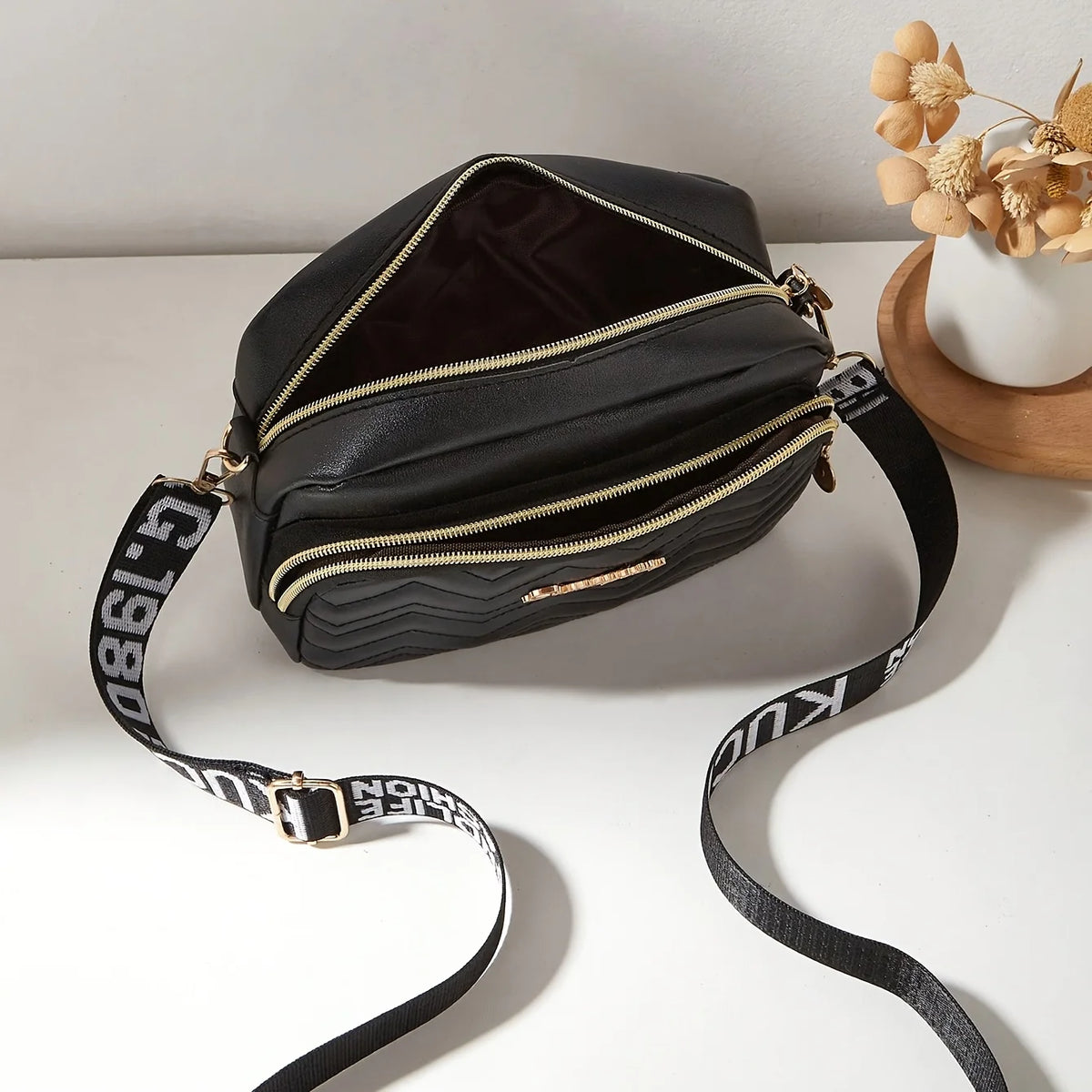Black quilted crossbody camera bag with a gold letter strap resting on a white surface.