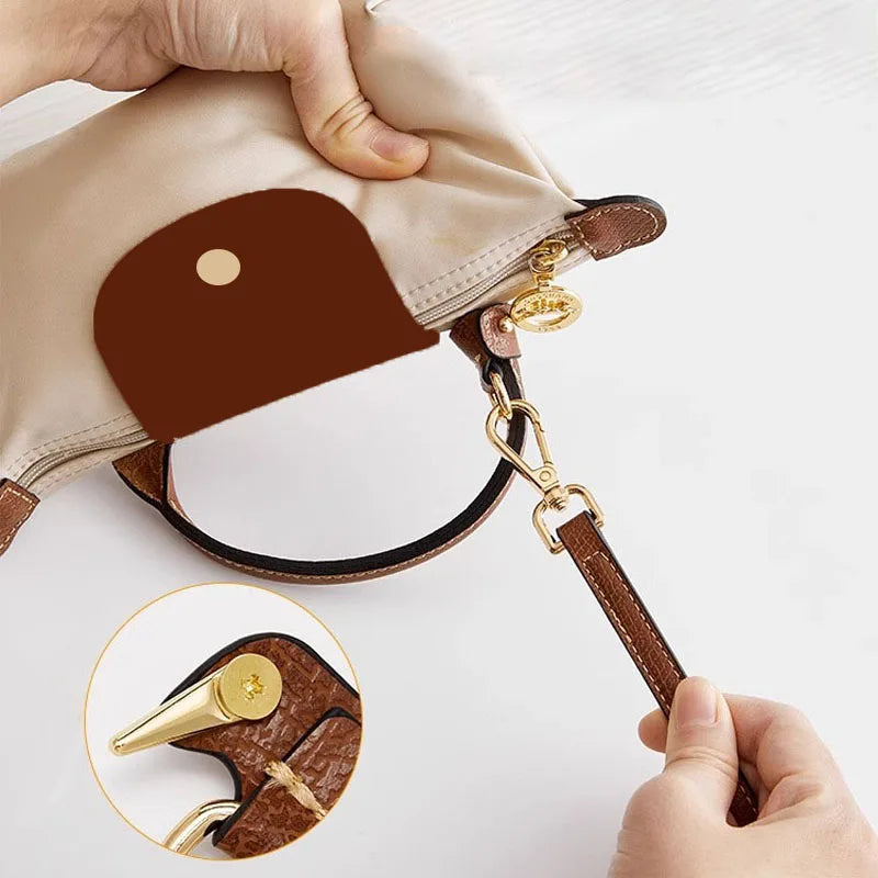A hand holds a tan leather crossbody strap with a brown leather pouch and gold hardware.