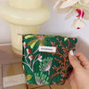 A green fabric cosmetic pouch with floral embroidery is held against a light background.