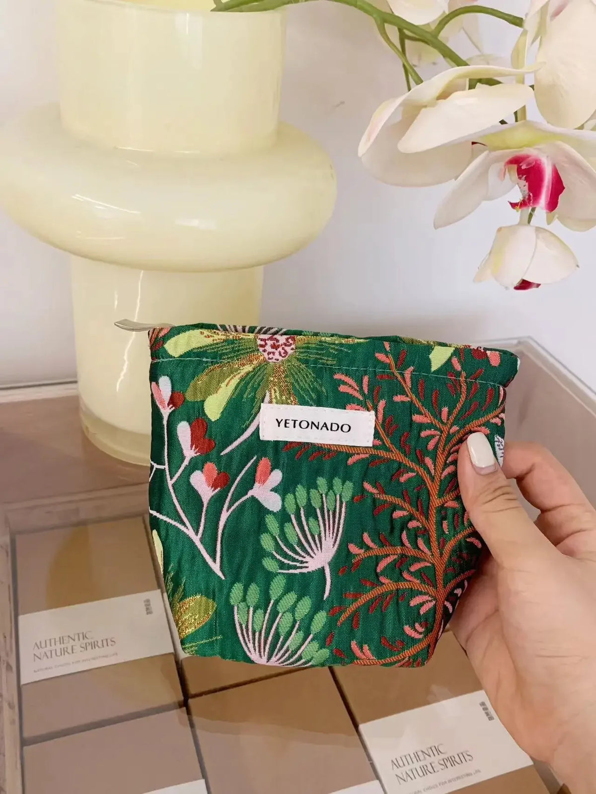 A green fabric cosmetic pouch with floral embroidery is held against a light background.