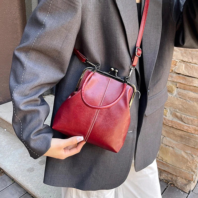 A woman holds a red leather shell handbag with a brown crossbody strap against a gray blazer.