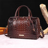 A dark brown crocodile pattern leather crossbody tote bag resting on a light surface next to a piece of wood.