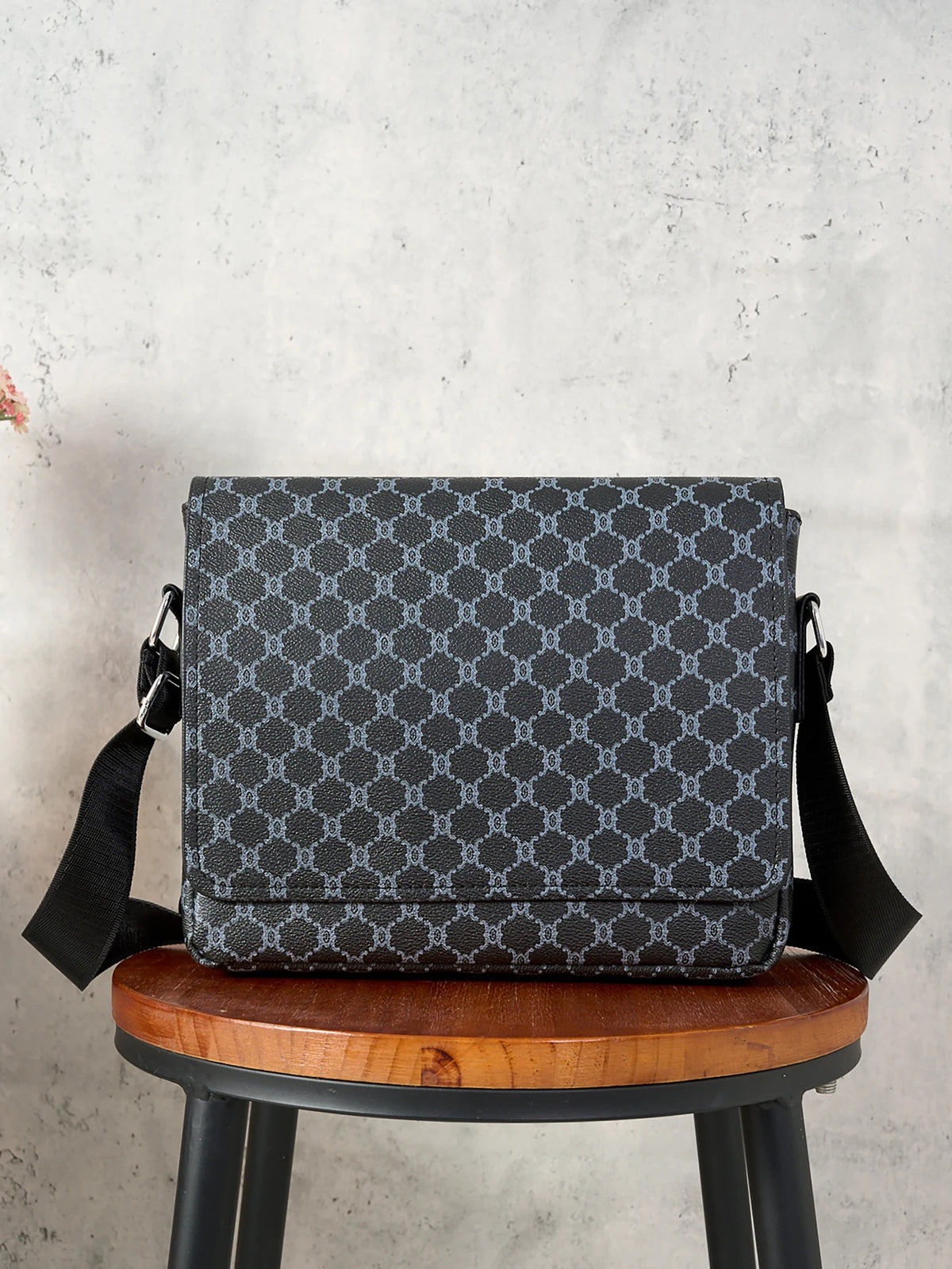 A dark gray patterned flap crossbody bag resting on a wooden stool against a light gray background.
