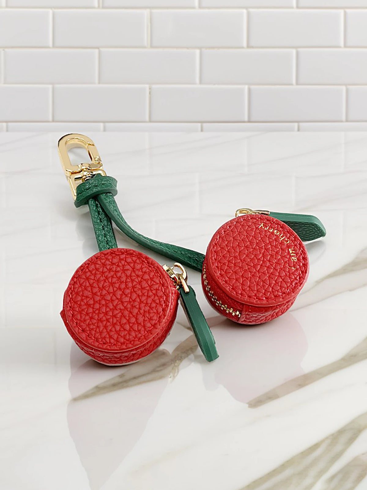 A red cherry fruit shape coin purse keychain with green leather strap resting on a white marble surface.