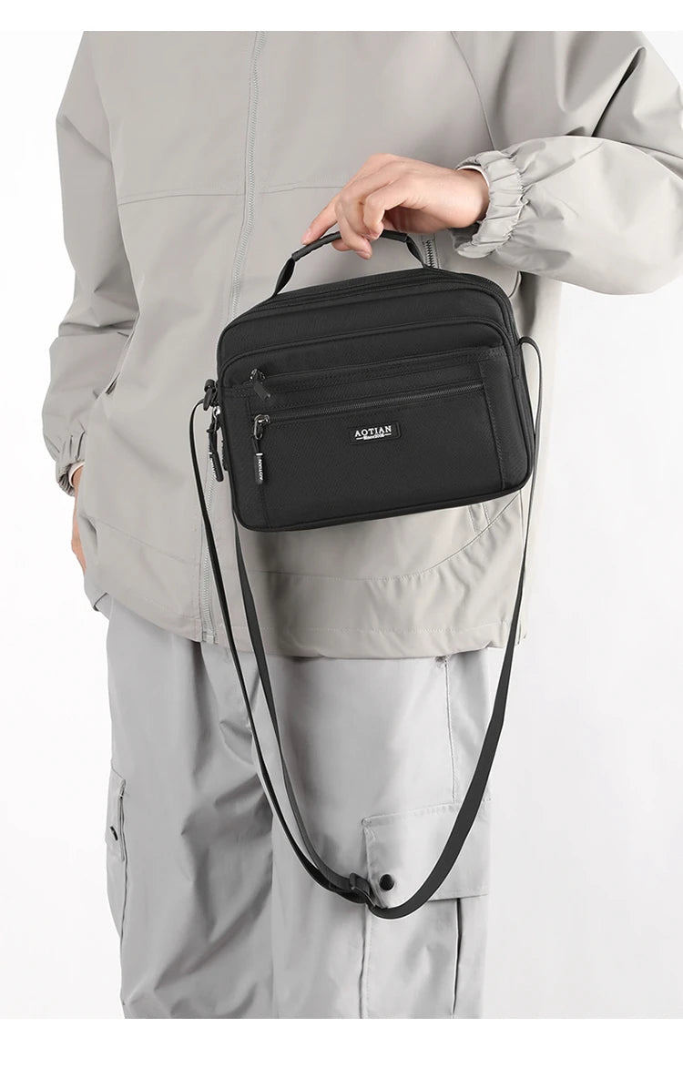 A hand holds a black compact waterproof crossbody messenger bag against a light gray jacket.