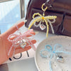 A hand holds a pink pearl bowknot keychain with a silver heart charm next to a dark brown leather bag.