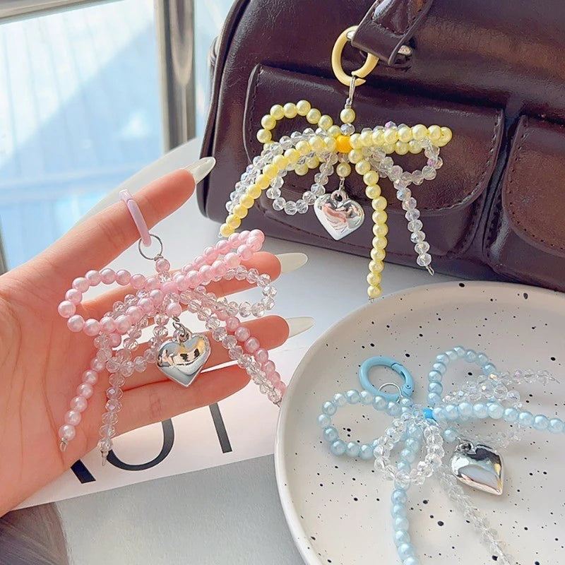 A hand holds a pink pearl bowknot keychain with a silver heart charm next to a dark brown leather bag.