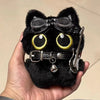 Colgante de llavero de un peluche de gato negro con grandes ojos amarillos y una hebilla plateada, sostenido en una mano.
