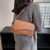 Una mujer con suéter gris sostiene un bolso cruz de cuero marrón estilo vintage contra un estante oscuro.