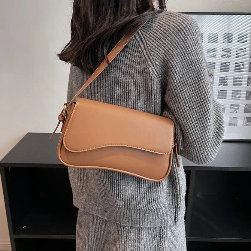 Una mujer con suéter gris sostiene un bolso cruz de cuero marrón estilo vintage contra un estante oscuro.