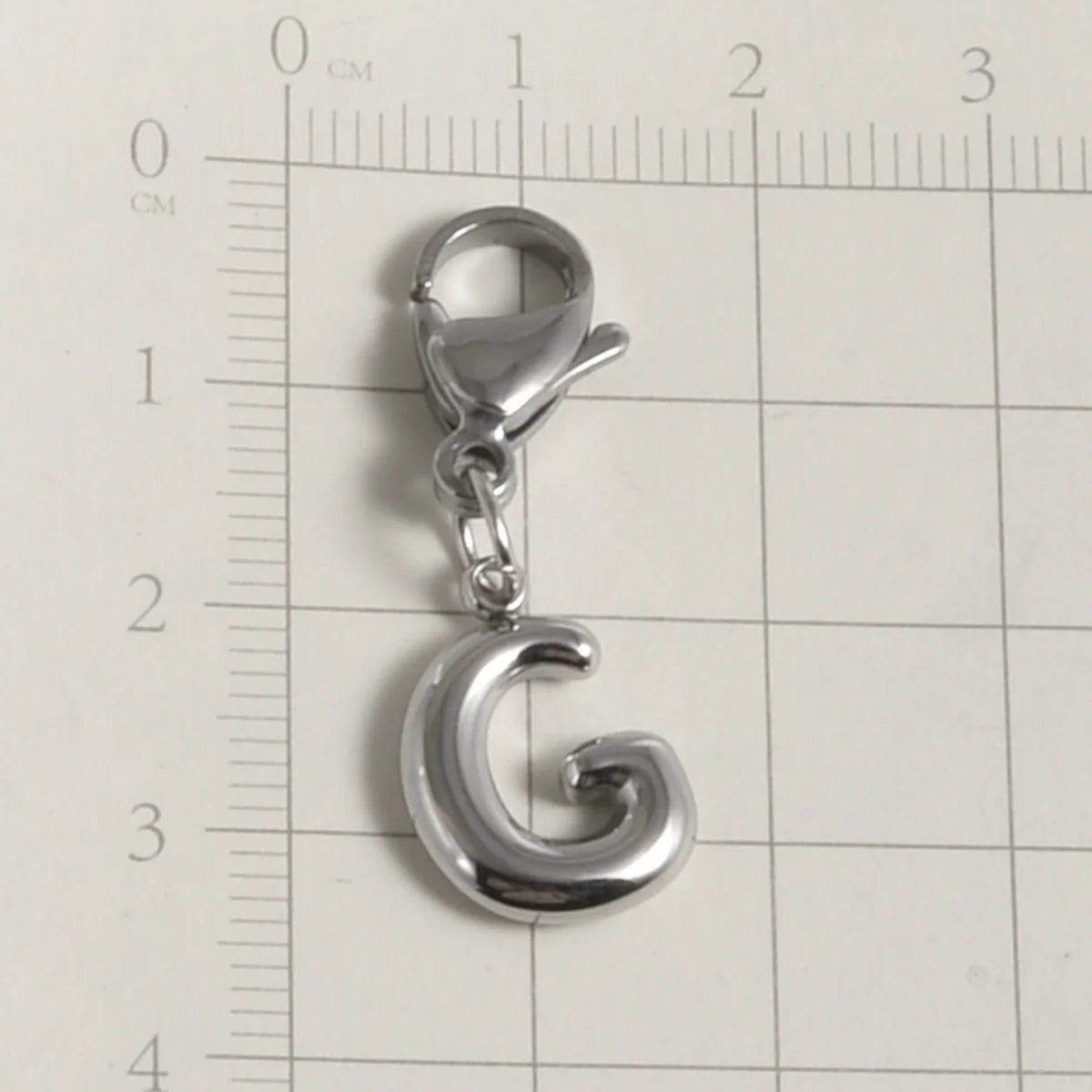 Silver stainless steel alphabet letter charm shaped like the letter G with a lobster clasp, measured against a ruler.