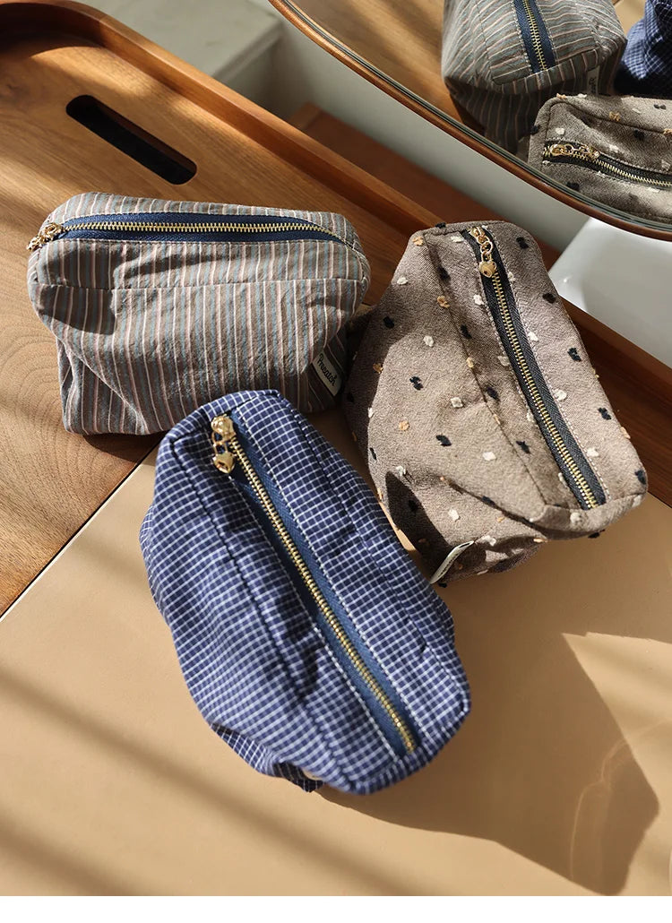 Trois pochettes de rangement cosmétique en tissu à motifs carreaux gris, marron et bleu sont présentées sur une surface en bo