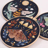 A dark embroidery kit featuring illustrations of a bee and butterflies on a circular fabric piece.
