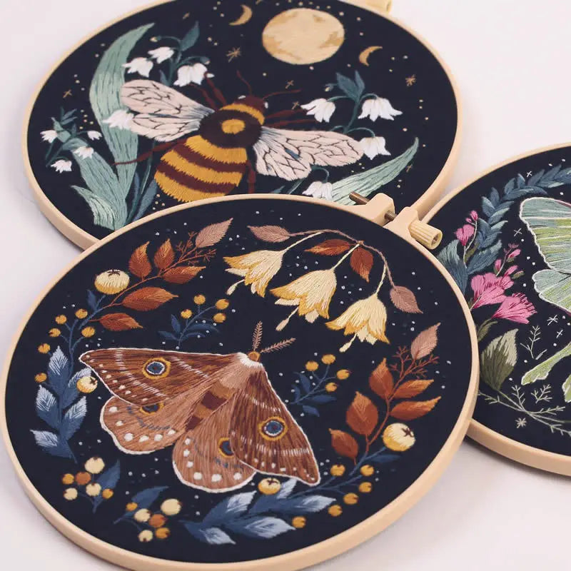 A dark embroidery kit featuring illustrations of a bee and butterflies on a circular fabric piece.