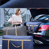 A woman stands next to a black car with open trunk, holding two navy blue storage bags.