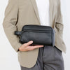 A black PU leather crossbody bag is held by a person wearing a beige blazer and gray pants.