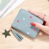 A light blue compact PU leather wallet with heart-shaped embellishments and a green tassel is held by a hand on a wooden surf