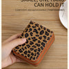 A hand holds a small leopard print wallet with an orange trim on a wooden surface.
