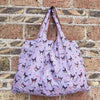 A purple reusable shopping bag featuring a pattern of small cats on a white background, displayed against a brick wall.