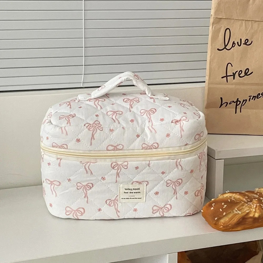 A white quilted cosmetic bag with pink embroidery rests on a shelf next to a brown paper bag.