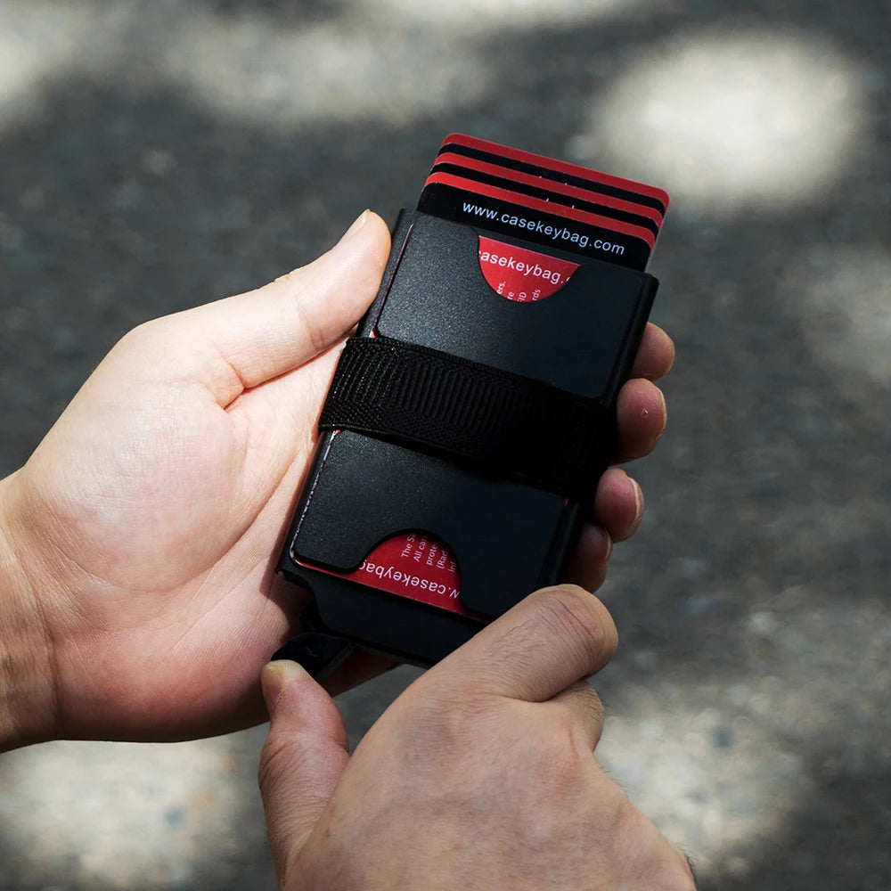 A hand holds a black RFID blocking metal pop-up card wallet with red accents and a black sleeve.