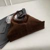 A brown suede slouchy shoulder bag resting on a white surface next to a gray blazer and a clear glass.