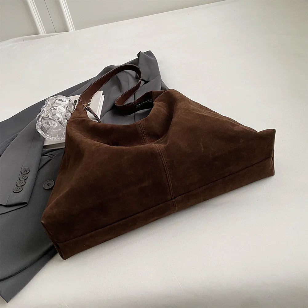 A brown suede slouchy shoulder bag resting on a white surface next to a gray blazer and a clear glass.