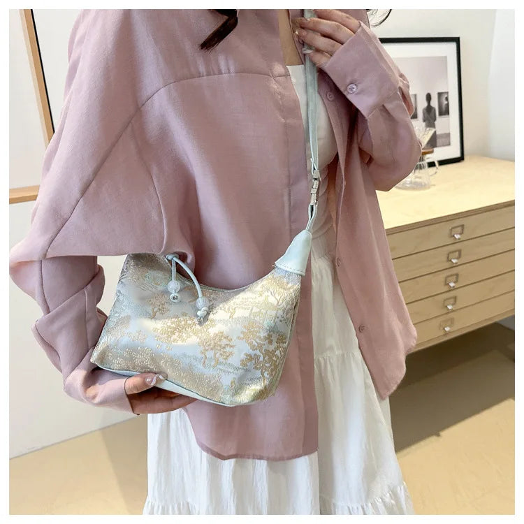 A woman holds a light pink jacket and a silver embroidered crossbody bag with gold accents.
