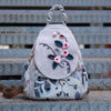 A light gray linen backpack featuring floral embroidery and a woven strap, resting on a wooden surface.